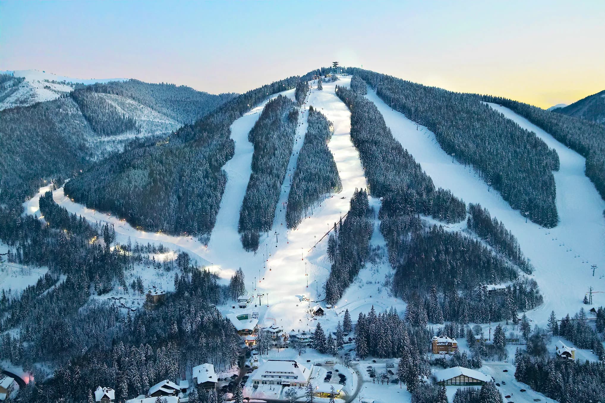 Verschneiter Hirschenkogel mit beleuchteten Skipisten und Gebäuden am Hangfuß